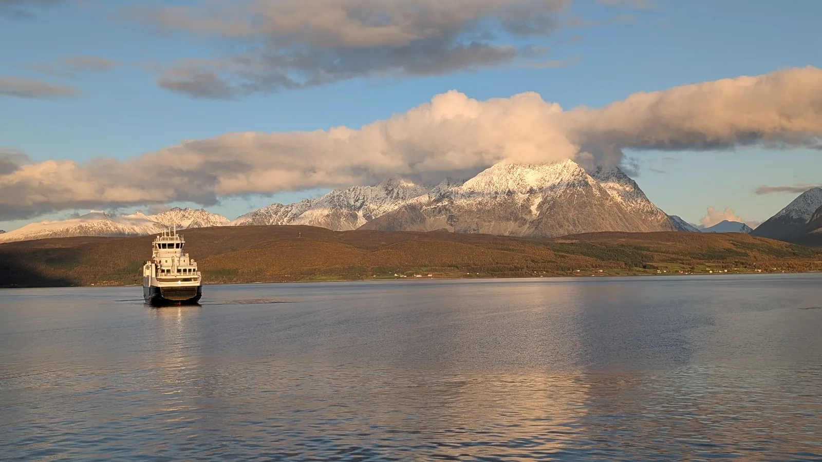 Golden evening light on a glassy sound: a small ferry passes beneath a brown mountain capped with snow.