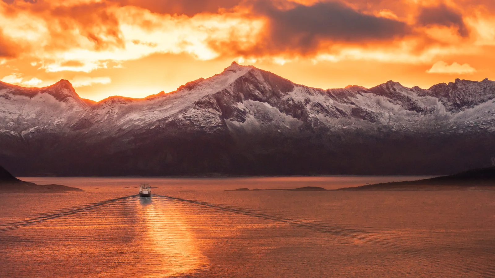 Blazing sunset over the Ullsfjord: orange clouds above snow-covered peaks, and the Brensholmen–Botnhamn ferry drawing its wake across golden water.