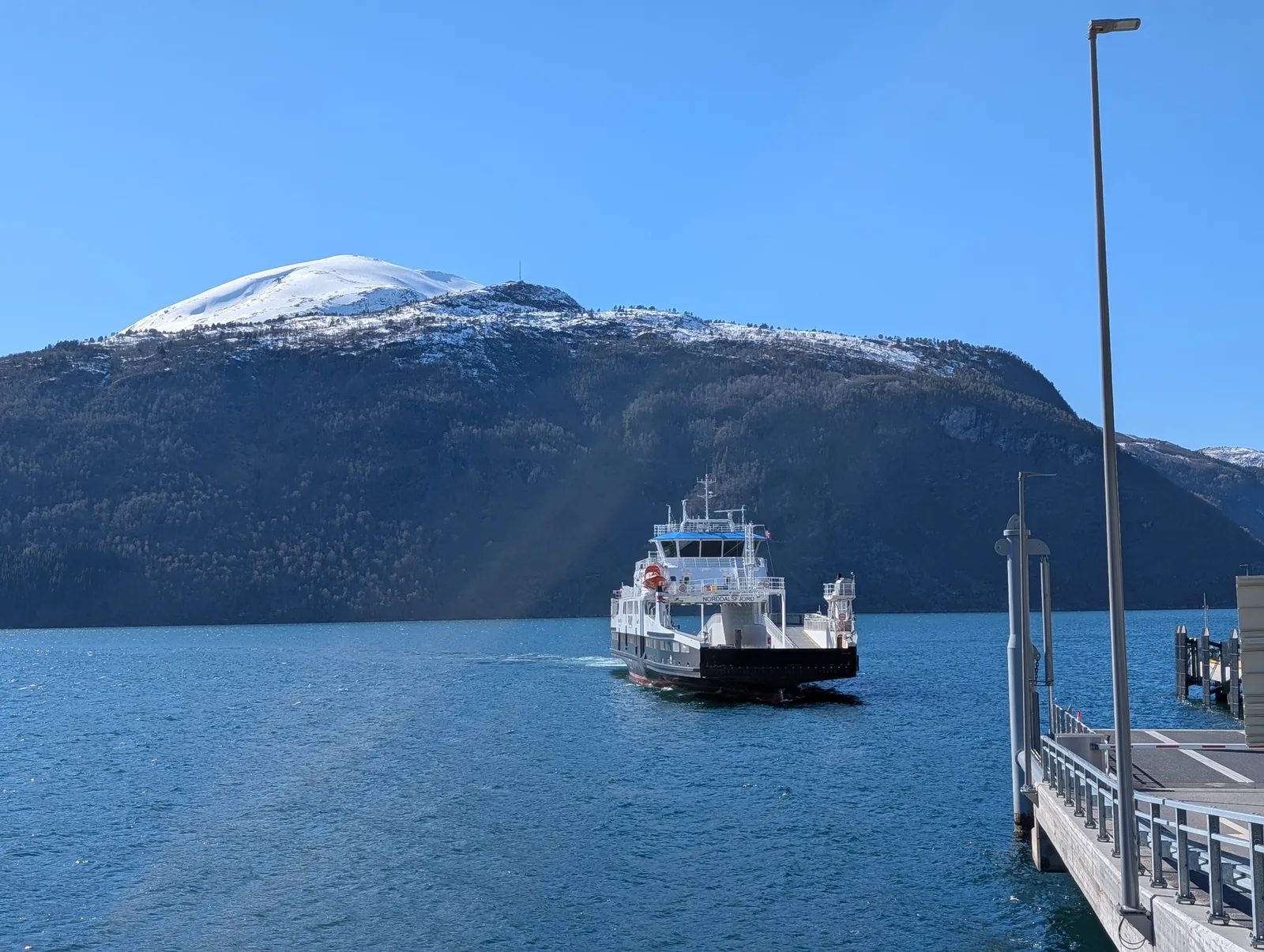 A white and red ferry approaching the slipway, a rounded mountain still under snow behind it and a deep blue sky.