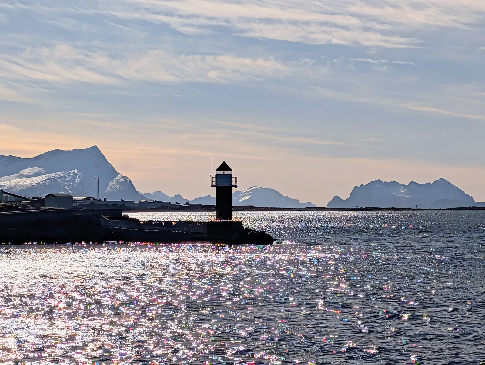 Backlit sparkling sea: the silhouette of a small lighthouse at the end of a pier and, far off, a line of jagged peaks.