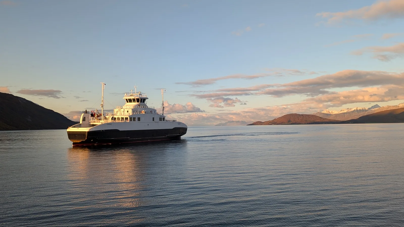 Backlit in golden evening light, a white ferry with a dark hull glides over glassy water.