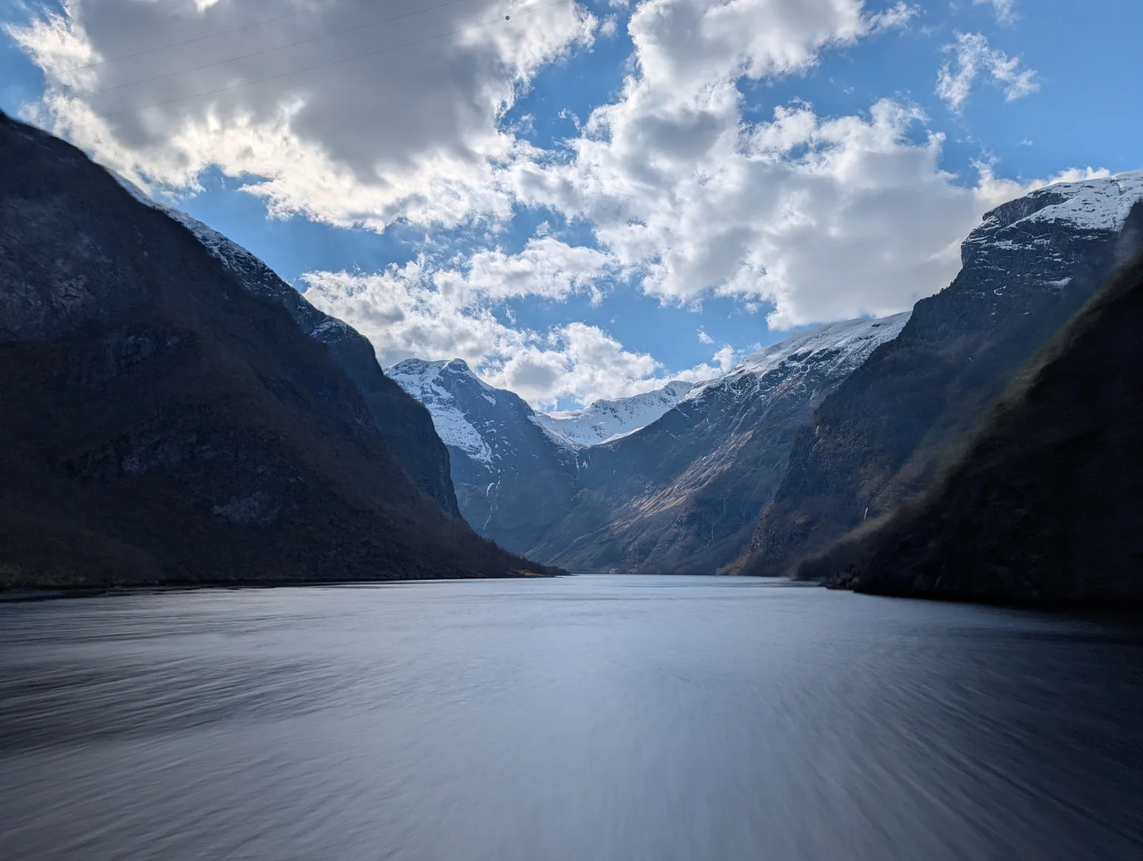 A narrow fjord with steep dark walls, mirror-flat water and patches of snow high on the slopes.