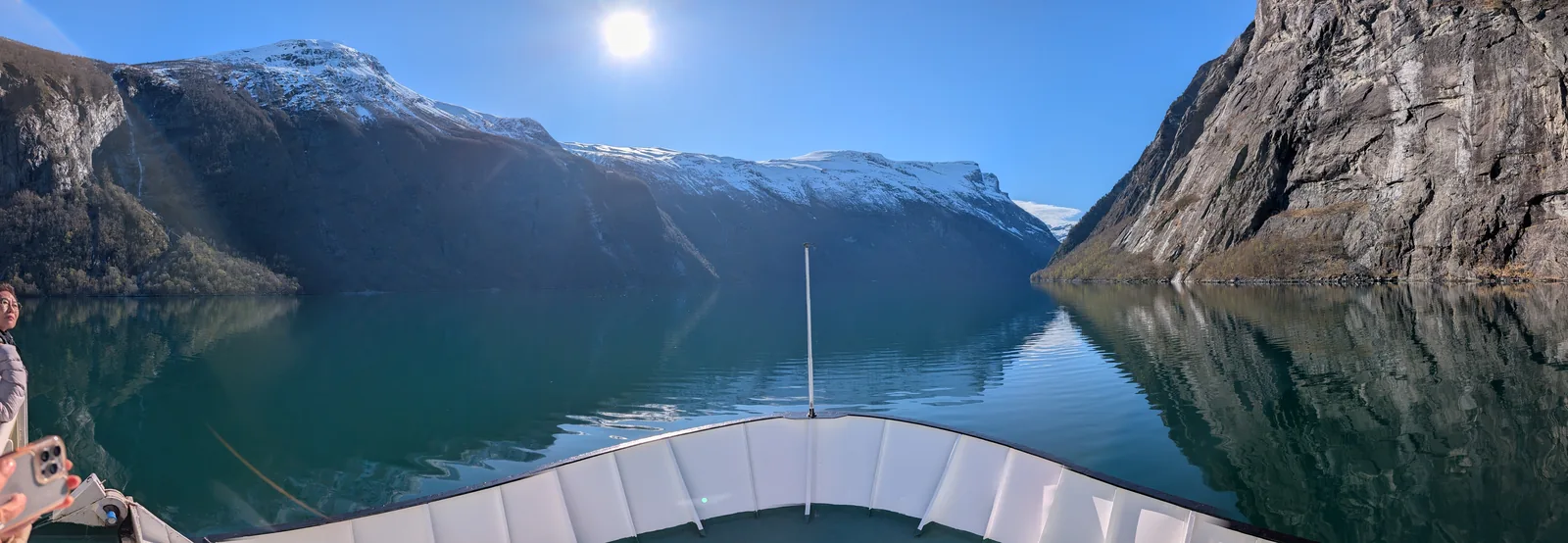 From a ferry's bow, a perfectly still fjord hemmed in by brown walls and snow-covered summits under a high sun.