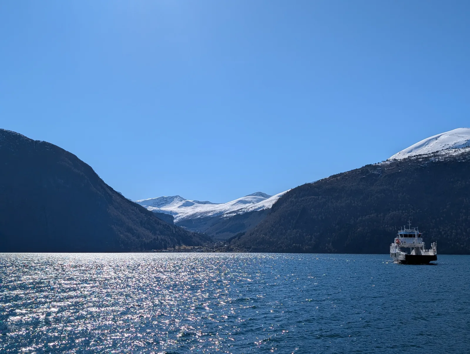 A small ferry far off on sparkling water between snow-covered mountains.