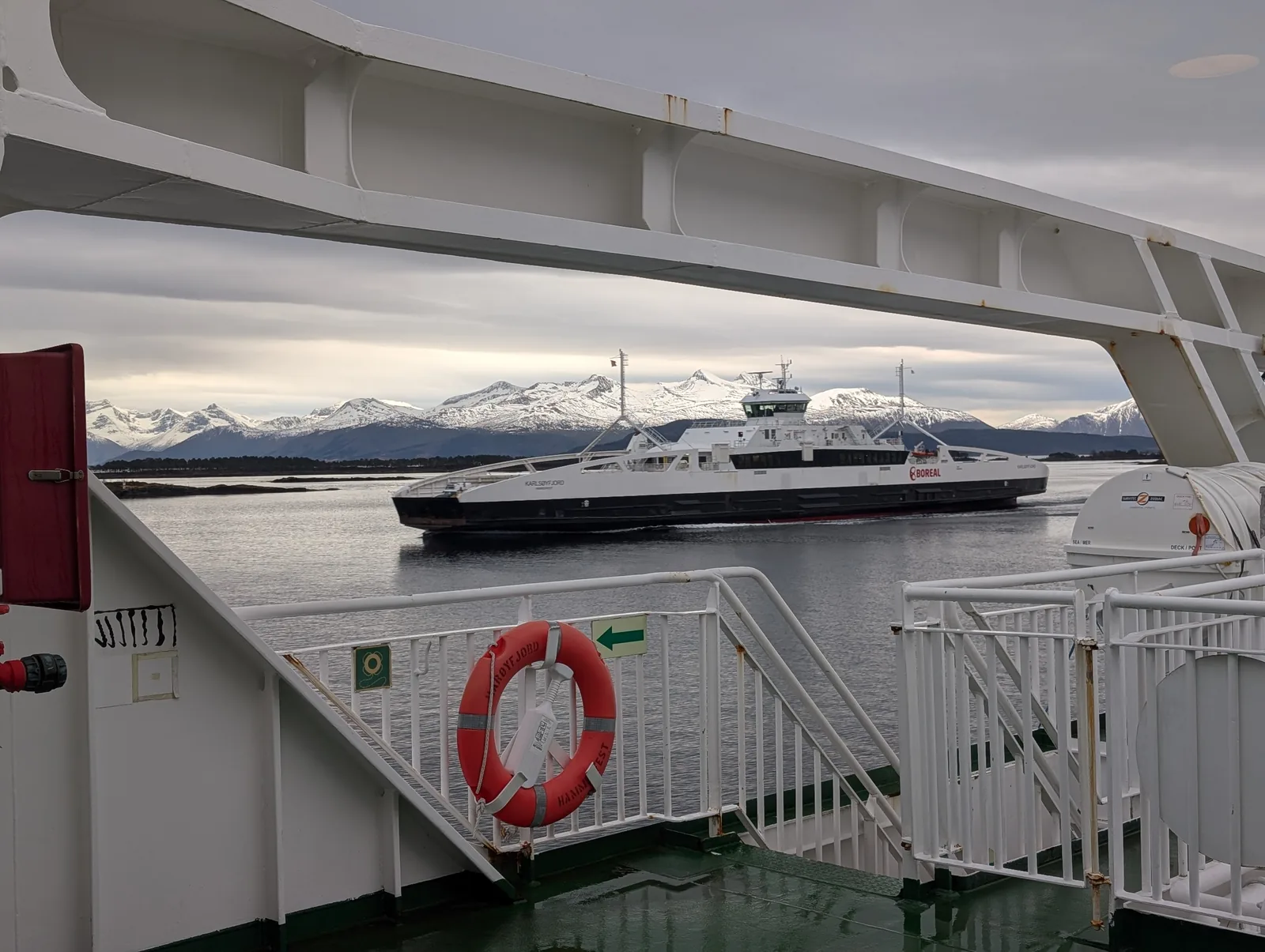 From a ferry's deck, a lifebuoy in the foreground and another ferry passing in front of snow-covered mountains.
