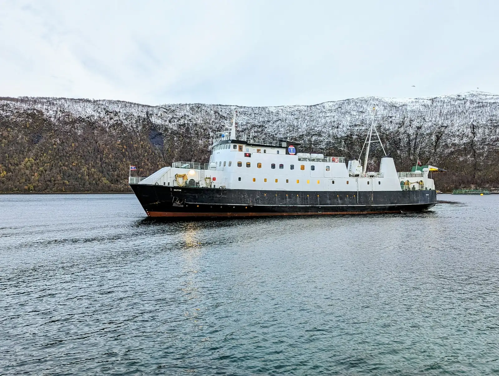 A white ferry with a dark hull glides over green-blue water in front of a snowy ridge and brown slopes.